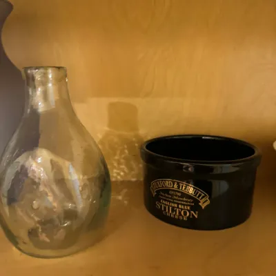 Small clear glass carafe paired with a Tuxford & Tebbutt English Blue Stilton Cheese black ceramic pot, displayed on a wooden shelf.