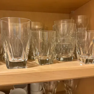 Collection of clear crystal glassware featuring faceted geometric bases, stacked on wooden cabinet shelves alongside white china cups.