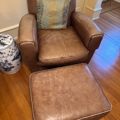 Distressed tan leather club armchair and matching ottoman with piping trim, accompanied by an embroidered accent pillow.