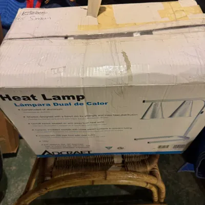Adcraft dual shade aluminum heat lamp, boxed in original retail packaging, resting on a woven bamboo footstool.