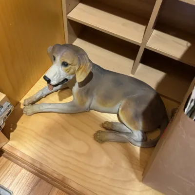 Realistic resin or painted ceramic statue of a lying hound dog with a tongue sticking out, resting on a wooden floor inside a cabinet.