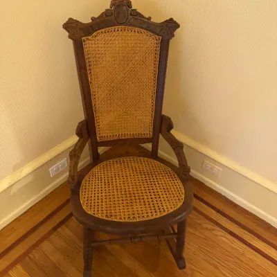 Antique carved wooden rocking chair featuring woven cane back panel and seat, decorated with ornate crest detailing.