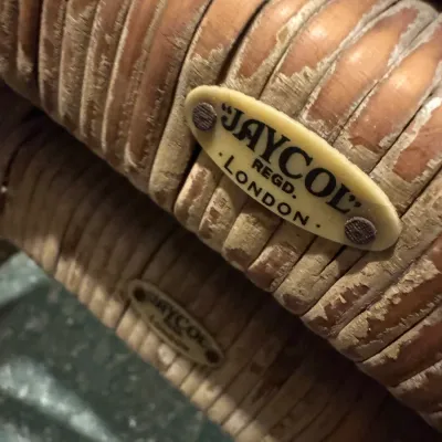Close-up detail of a Jaycol London maker's mark plaque attached to a vintage coiled leather or wicker handle, displaying wear and...