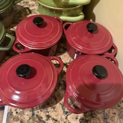 Set of four mini burgundy ceramic cocottes with lids, displayed on a granite countertop next to stacked green bakeware.