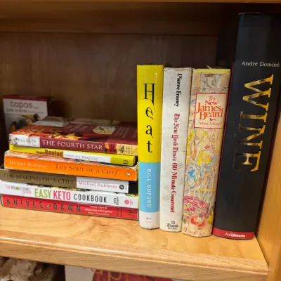 Collection of culinary and food books stacked and standing on a light wood bookshelf, featuring titles like Heat by Bill Buford and Wine...