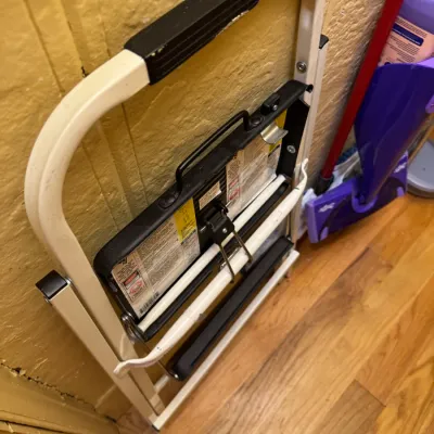 Folded white and black metal two-step stool leaning against a wall, showing safety labels and light cosmetic wear.