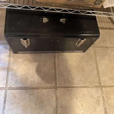 Vintage Craftsman black metal toolbox featuring metal draw latches and a top carry handle, stored beneath a wire shelf.