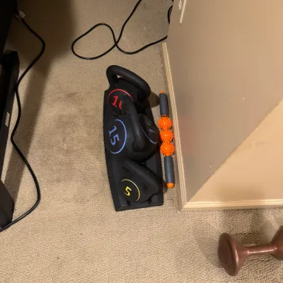 Set of three color-coded kettlebells on a base rack, including 5 lb, 10 lb, and 15 lb weights, alongside a massage roller stick.