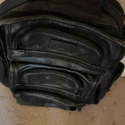 Black Tumi leather backpack featuring multiple zippered exterior compartments and a top carry handle, showing some minor wear.