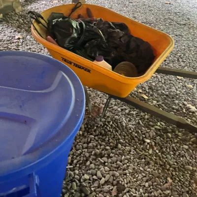 True Temper yellow plastic wheelbarrow filled with assorted yard tools, seated next to a blue plastic storage container outdoors.