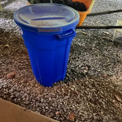 Blue plastic trash bin with lid placed on a gravel surface alongside an orange wheelbarrow.