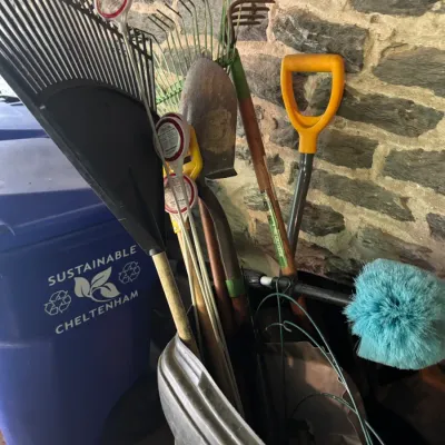 Collection of long-handled yard tools including leaf rakes, a metal shovel, garden cultivator, and duster stored in a bin against a stone...