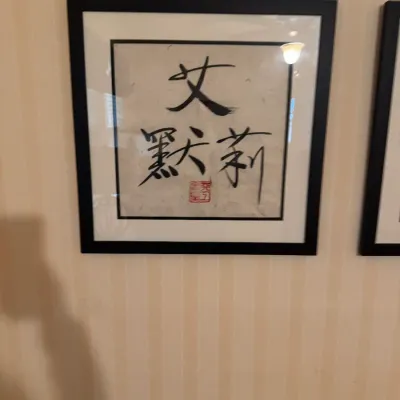 Framed East Asian calligraphy art piece featuring black ink characters and a red artist seal, presented in a black frame with white matting.