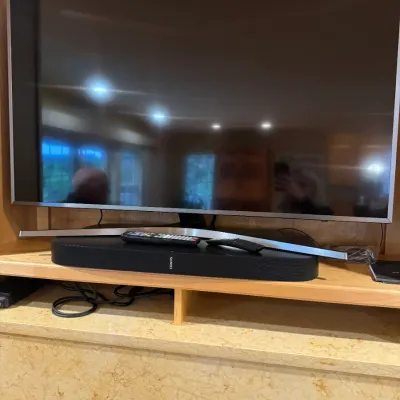 Samsung flat-screen television paired with a black Sonos soundbar and remotes, displayed on a wooden shelf over a fireplace mantle.