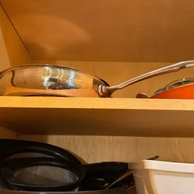 Stainless steel cookware and kitchen utensils displayed on wooden cabinet shelves, including a fry pan and fine mesh strainers.