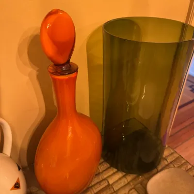 Mid-century modern vibrant orange glass decanter with stopper standing alongside a tall olive green glass cylindrical vase.