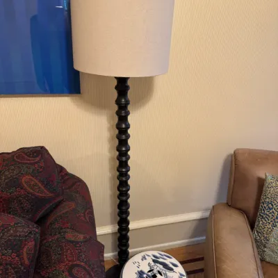 Tall floor lamp featuring a black turned-wood spindle stem and a beige fabric drum shade, positioned between seating.