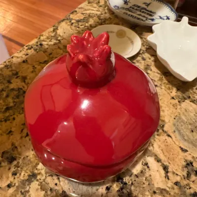 Glossy red ceramic pomegranate-shaped lidded dish or candy jar, shown alongside small decorative porcelain dishes on a granite countertop.