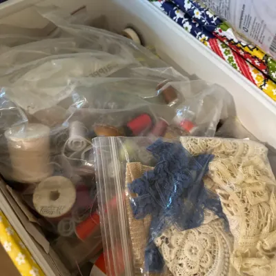 Collection of sewing notions including spools of thread, lace trim, and fabric remnants stored in plastic bags inside a sewing box.