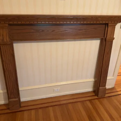 Traditional stain-finished wooden fireplace mantel with dentil molding trim and fluted side columns, set against a striped wallpapered wall.