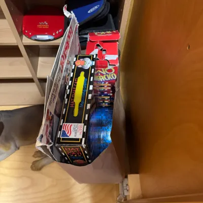 Paper bag filled with vintage board games, including Twister and Uno Rack-O, sitting next to a shelf with compact discs and a red Sony...