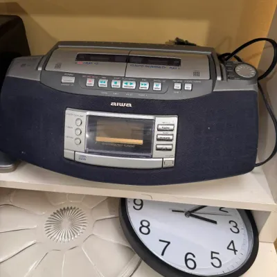 Vintage Aiwa portable CD player and cassette boombox resting on a shelf, displayed with a round wall clock below.