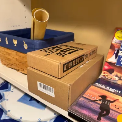 Shelf filled with Judaica items, including a woven basket with embroidered blue lining, boxed sets with Hebrew lettering, a Rick Recht...