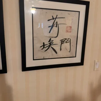 Framed Asian calligraphy art featuring black ink characters and a red artist stamp on textured fibrous paper, presented in a black wood...