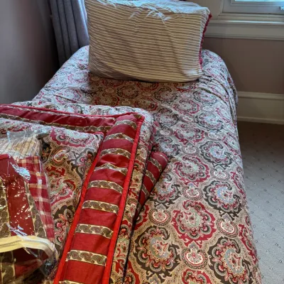 Twin bed outfitted with a red and beige paisley printed comforter set, complete with matching throw pillows and additional packaged bedding.