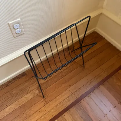 Mid-century modern black wire magazine rack resting on hardwood flooring.