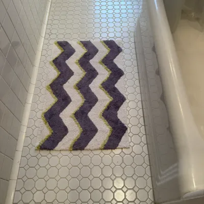 Modern plush bath mat featuring a chevron pattern in purple, lime green, and white, displayed on a tiled bathroom floor next to a white...