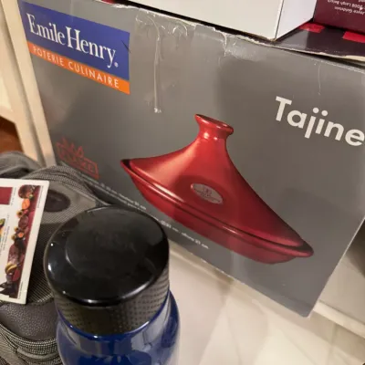 Boxed Emile Henry red ceramic tajine, accompanied by a blue water bottle and small travel pouch on a shelf.