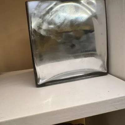 Square clear glass art piece or decorative block with textured inner detail, resting on a white shelf.