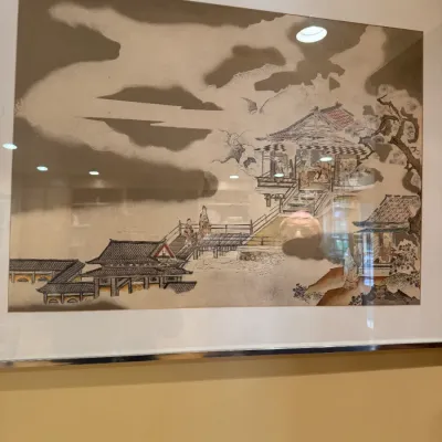 Framed Japanese-style landscape artwork featuring traditional architectural elements and figures, presented in a silver-toned frame under...