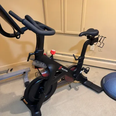 Peloton stationary exercise bike with attached touchscreen monitor, red resistance knob, and rear weight racks, shown in good working...