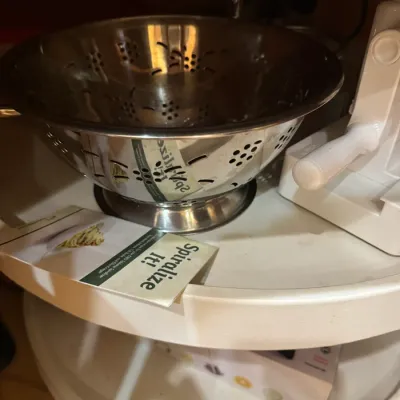 Stainless steel footed colander sitting on a lazy Susan shelf alongside a white plastic vegetable spiralizer with its instruction booklet.