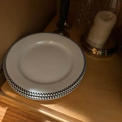 Stack of white ceramic plates with a black and white checkered trim pattern, displayed on a wooden shelf.