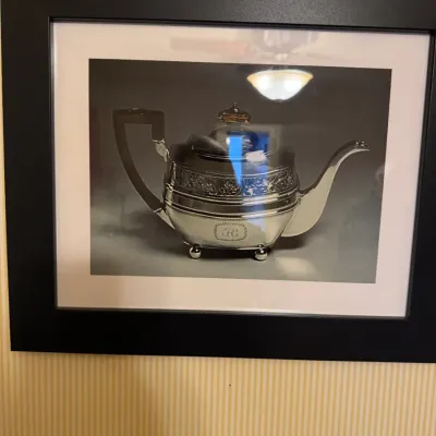 Framed black and white photograph depicting an ornate antique silver teapot, set within a wide white mat and a thick black frame.