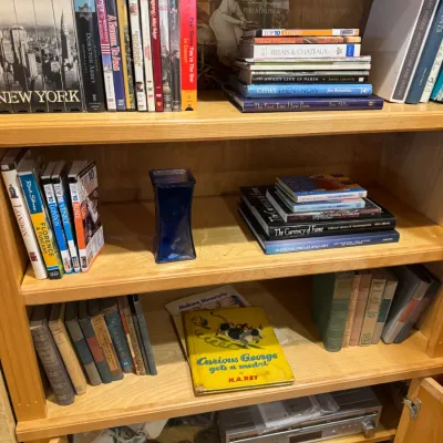 Wooden bookcase filled with mixed media, featuring a vintage Curious George children's book, various art and travel hardcovers,...