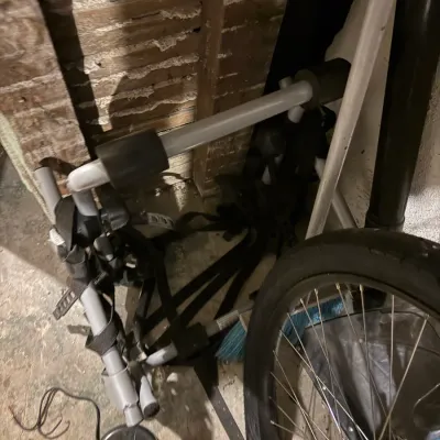 Trunk-mounted vehicle bicycle rack with silver metal frame, black foam padding, and attachment straps, stored alongside a bicycle wheel.