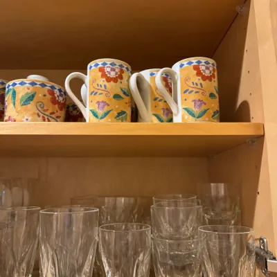 A collection of glassware and ceramic tableware displayed on wooden cabinet shelves, including colorful floral mugs and an assortment of...