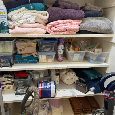 Linen closet shelves filled with folded towels, blankets, plastic storage bins, cleaning supplies, and vacuum cleaners.