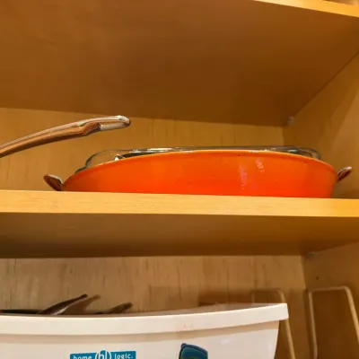 Orange enameled cast iron roasting pan with side handles and glass lid stored inside a wooden kitchen cabinet.