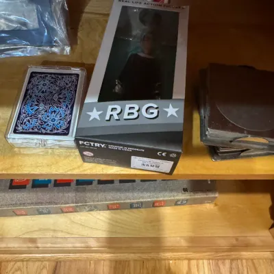 FCTRY Ruth Bader Ginsburg 'RBG' real life action figure in original packaging, shown on a wooden shelf alongside playing cards...