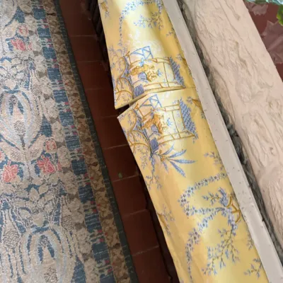 Roll of yellow chinoiserie wallpaper featuring blue scenic illustrations, shown alongside a vintage runner rug.