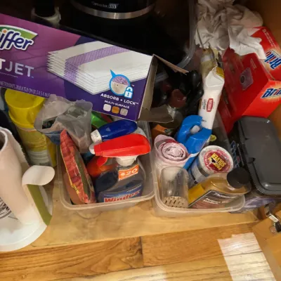 Assorted household cleaning supplies and tools stored in plastic bins under a cabinet, including Windex spray, Swiffer WetJet refills,...