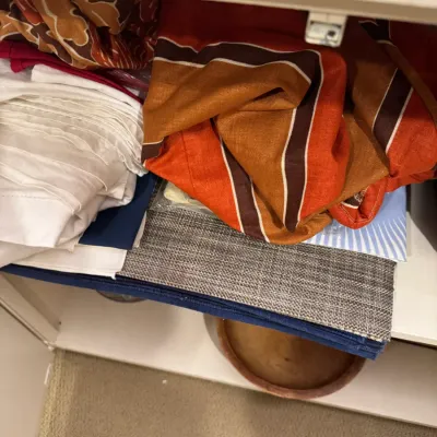 Assorted table linens and fabrics stored in a cabinet, featuring white cloth napkins, a woven grey placemat, and 1970s striped orange and...