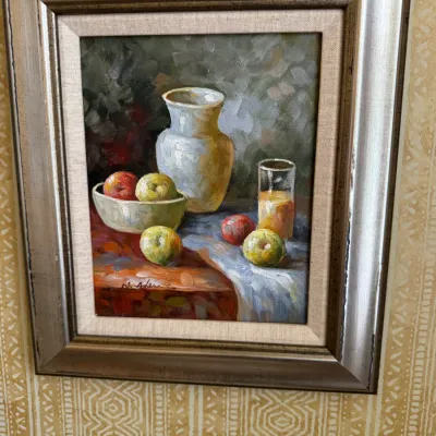 Framed still life oil painting depicting a ceramic pitcher, bowl of fruit, and a glass, housed in a metallic silver-toned frame with a...