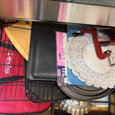 Collection of kitchen and baking supplies inside a drawer, including a red cast iron apple peeler, paper doilies, baking pans, a cooling...