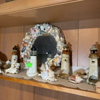 Sale Photo Thumbnail #124: Collection of nautical decor items including ceramic and painted lighthouse figurines, natural sea shells, and a seashell-encrusted oval mirror.